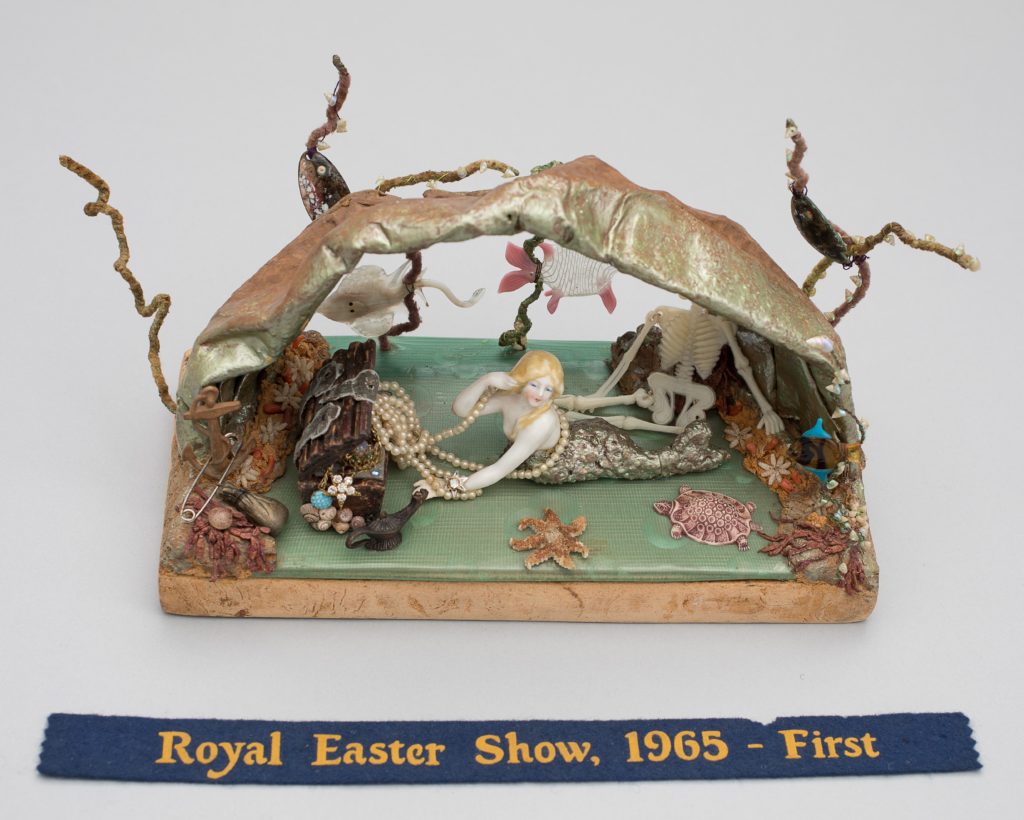 This doll tableau comprises an underwater cave with reclining ceramic mermaid with long peal necklace surrounded by marine props including a ceramic starfish and turtle, plastic skeleton and jewel-filled treasure chest. The tableau is made of various materials including beadwork, shells, plastic and pipe cleaners. The tableau is mounted on particle board. It has a blue ribbon "Royal Easter Show 1965 First".